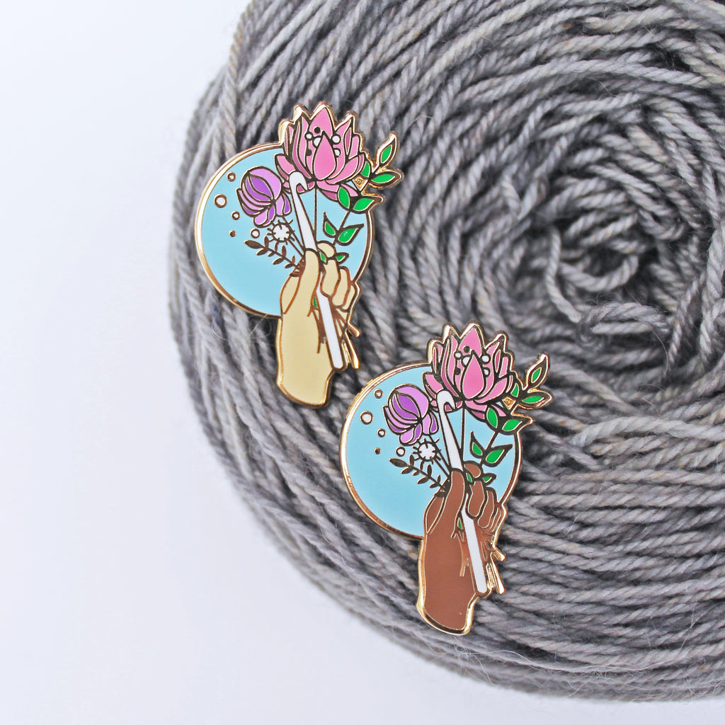 Floral enamel pin for crocheters. A crochet hook being held with a bouquet of flowers, available in multiple skin tones. 